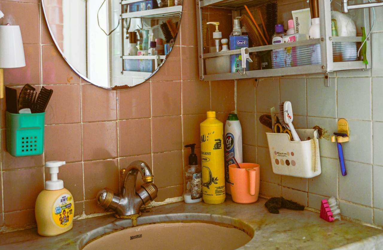 Un lavandino del bagno affollato con numerosi flaconi di plastica colorati, spazzolini da denti, pettini in contenitori a muro e uno specchio rotondo