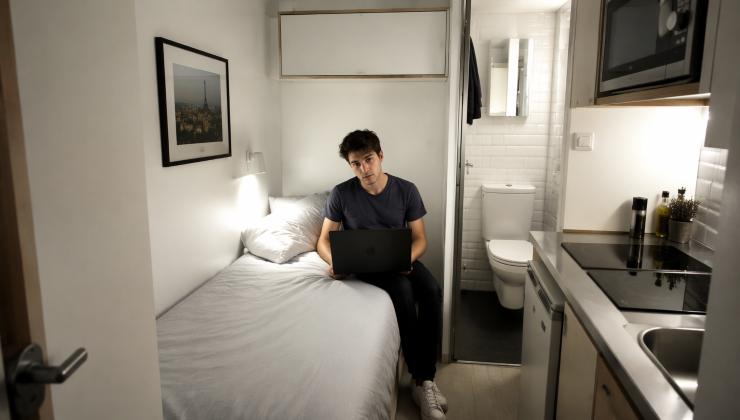 Uno studente fuori sede seduto su un letto singolo in un piccolissimo e moderno monolocale con cucina e bagno a vista nello stesso spazio
