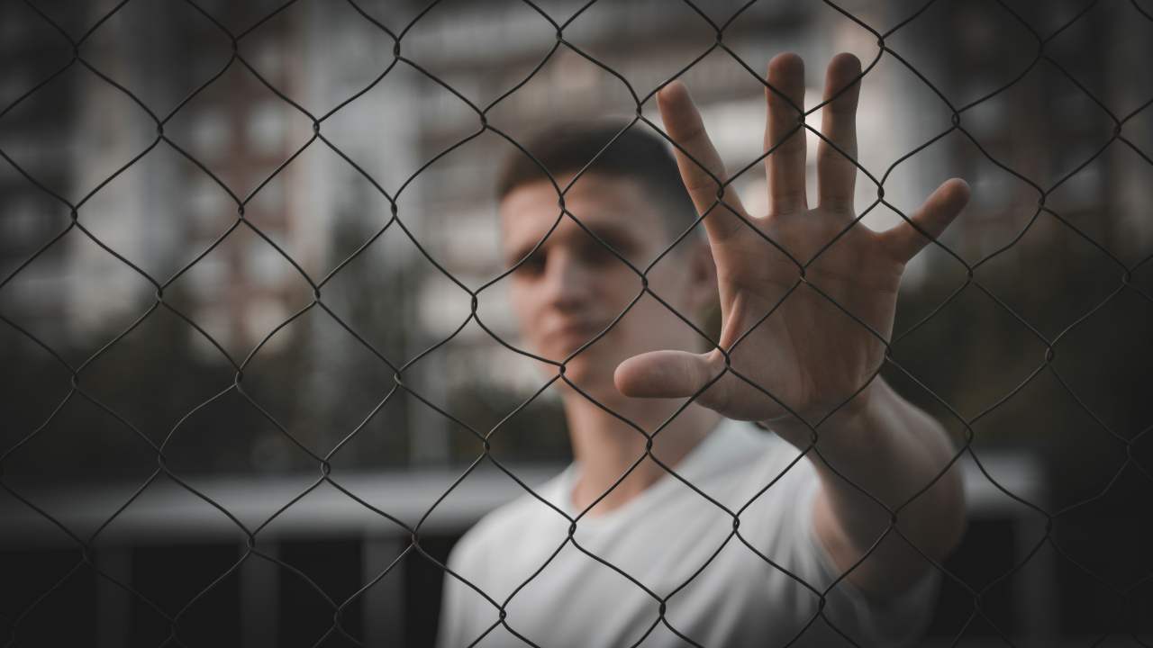 Un ragazzo sfocato dietro una rete metallica che poggia la mano sulla recinzione
