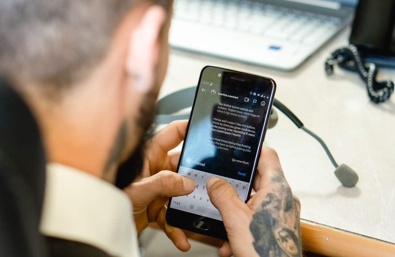 Uomo tatuato di spalle mentre scrive un messaggio sullo smartphone seduto alla scrivania con un computer portatile sullo sfondo